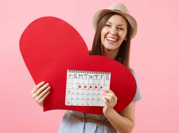 Eine glückliche Frau hält ihren Schwangerschaftskalender hoch, hinter dem sich ein großes rotes Papierherz befindet.