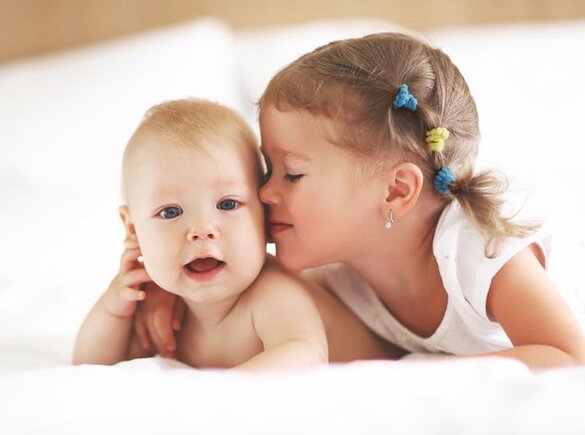 Eine Schwester gibt ihrem glücklichen kleinen Geschwisterchen einen Kuss auf die Wange, während sie auf dem Bauch in einem Bett liegen.