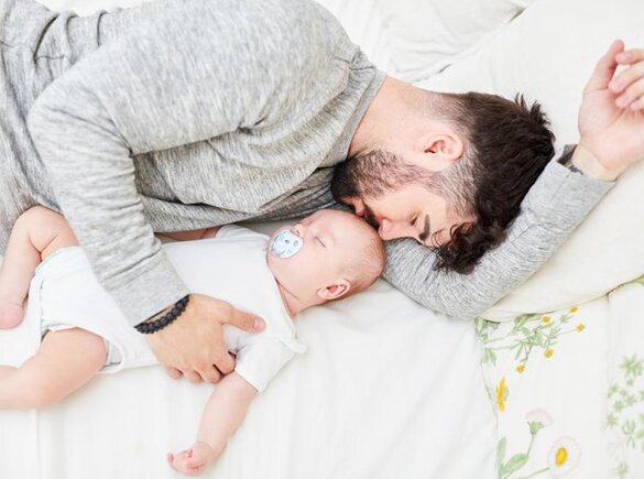Vater und Baby schlafen im Bett nebeneinander, der Vater hält das Baby.
