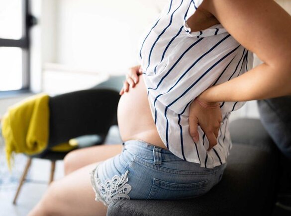 Eine schwangere Frau sitzt da und hält sich den Rücken und den schwangeren Bauch.