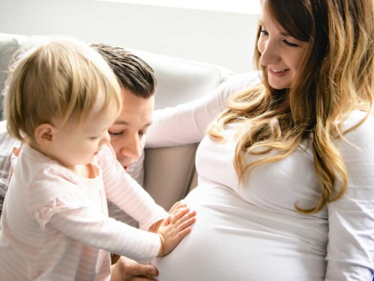 Eine Familie sitzt zusammen, während das erstgeborene Kleinkind mit dem Vater den schwangeren Bauch der Mutter berührt, während die Mutter es anlächelt.