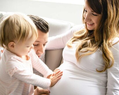 Eine Familie sitzt zusammen, während das erstgeborene Kleinkind mit dem Vater den schwangeren Bauch der Mutter berührt, während die Mutter es anlächelt.