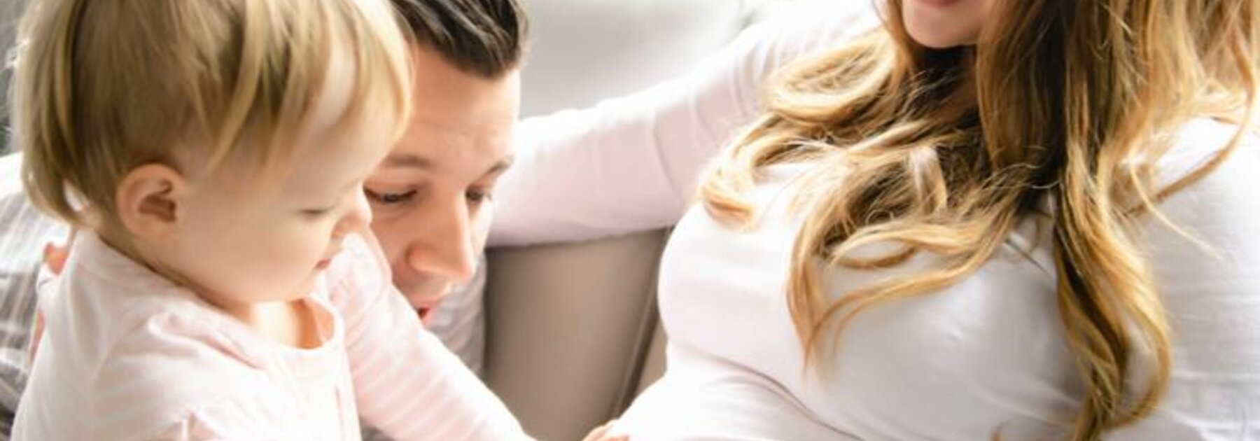 Eine Familie sitzt zusammen, während das erstgeborene Kleinkind mit dem Vater den schwangeren Bauch der Mutter berührt, während die Mutter es anlächelt.