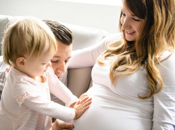 Eine Familie sitzt zusammen, während das erstgeborene Kleinkind mit dem Vater den schwangeren Bauch der Mutter berührt, während die Mutter es anlächelt.
