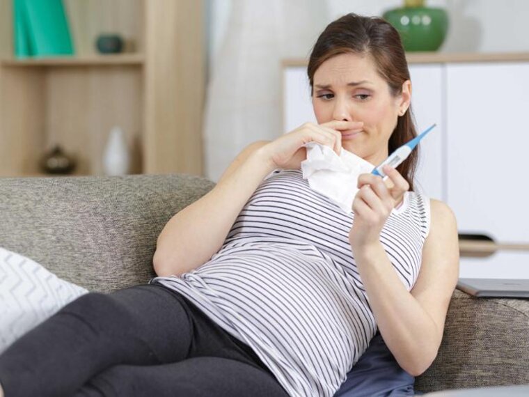 Eine schwangere Frau fühlt sich nicht wohl.  Sie liegt auf der Couch und hält ein Tuch und ein Thermometer in der Hand.