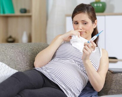 Eine schwangere Frau fühlt sich nicht wohl.  Sie liegt auf der Couch und hält ein Tuch und ein Thermometer in der Hand.