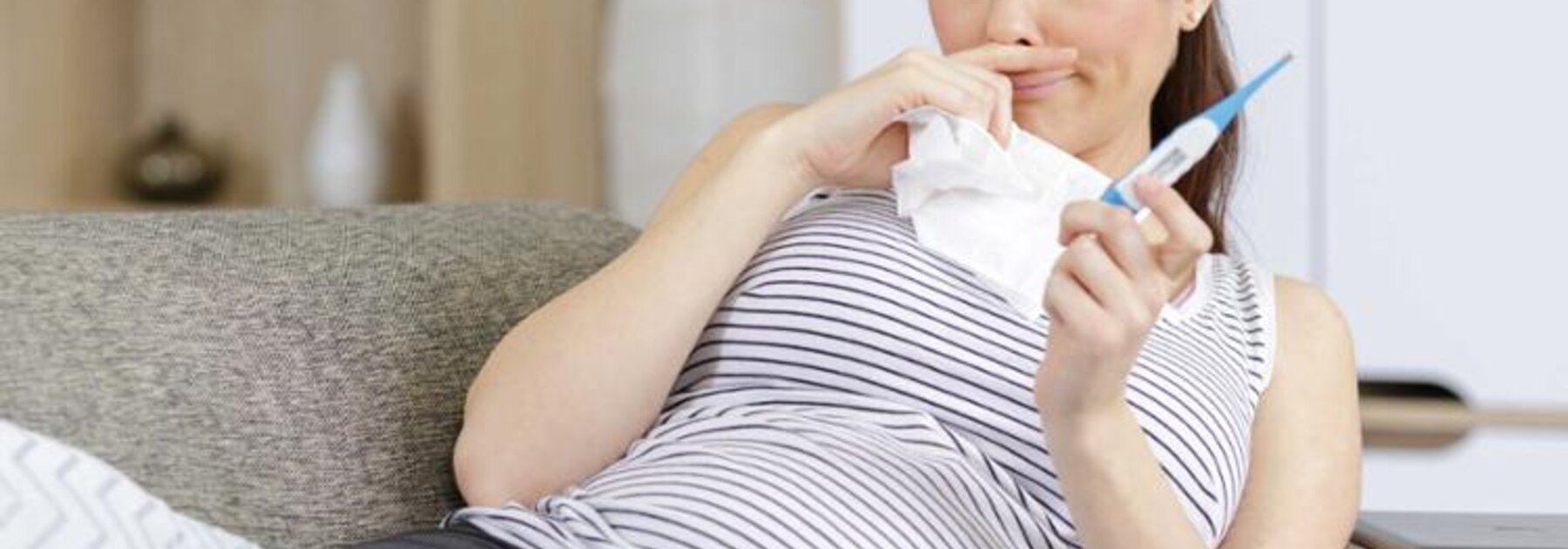 Eine schwangere Frau fühlt sich nicht wohl.  Sie liegt auf der Couch und hält ein Tuch und ein Thermometer in der Hand.