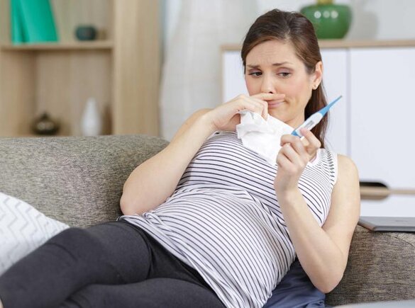 Eine schwangere Frau fühlt sich nicht wohl.  Sie liegt auf der Couch und hält ein Tuch und ein Thermometer in der Hand.
