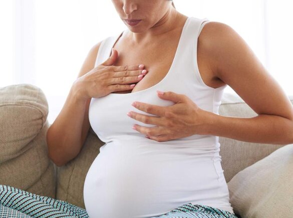 A pregnant woman massages her sore breast while sitting on a couch.