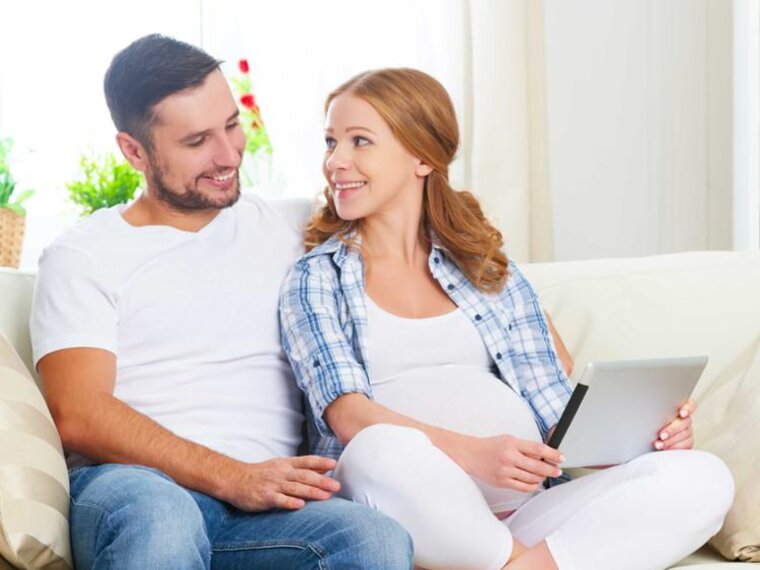 Schwangere Frau sitzt auf der Couch neben ihrem Mann und schaut auf ein Tablet.