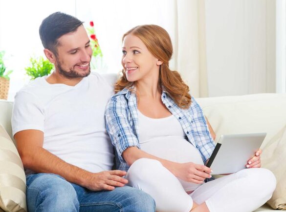 Schwangere Frau sitzt auf der Couch neben ihrem Mann und schaut auf ein Tablet.