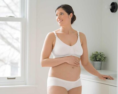 Eine schwangere Frau steht in ihrem Schlafzimmer und trägt einen Medela Keep Cool™ Ultra Komfort Atmungsaktiver Schwangerschafts- und Still-BH.