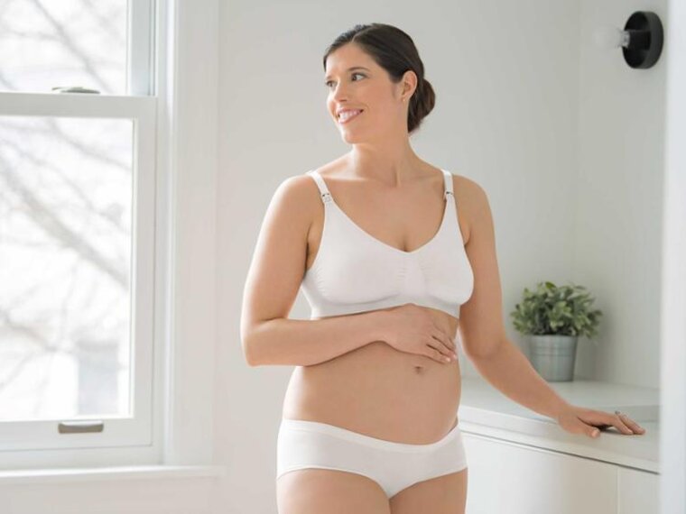 Eine schwangere Frau steht in ihrem Schlafzimmer und trägt einen Medela Keep Cool™ Ultra Komfort Atmungsaktiver Schwangerschafts- und Still-BH.