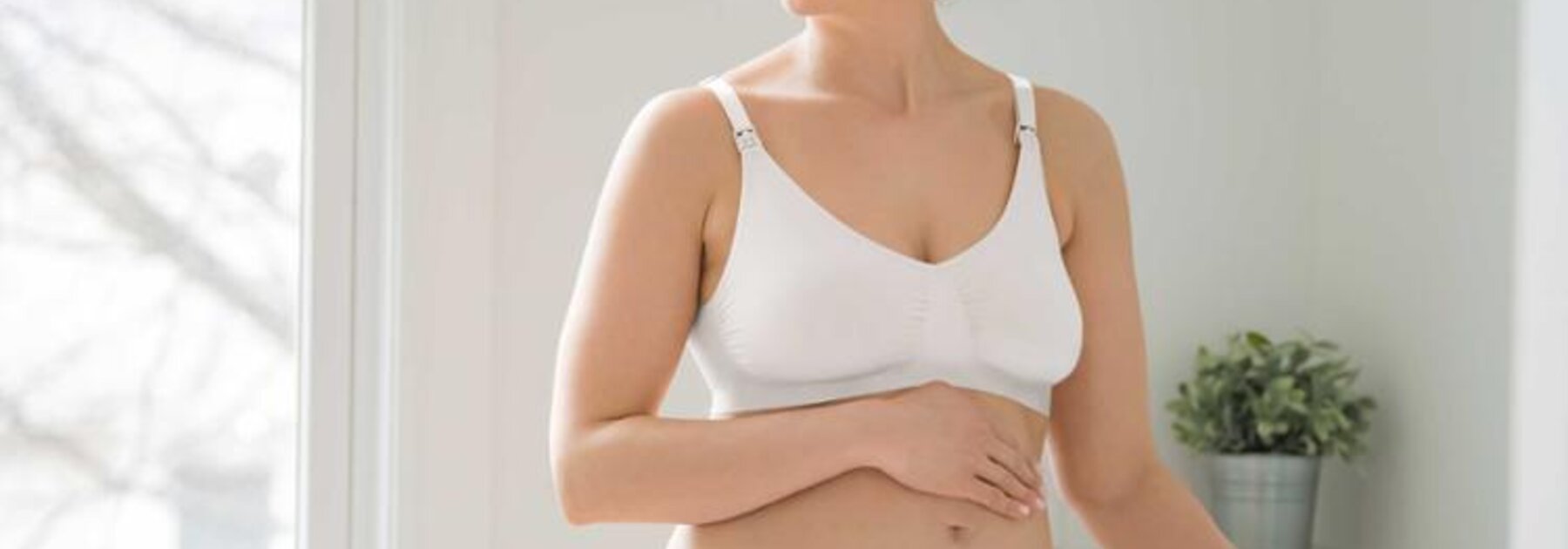 Eine schwangere Frau steht in ihrem Schlafzimmer und trägt einen Medela Keep Cool™ Ultra Komfort Atmungsaktiver Schwangerschafts- und Still-BH.