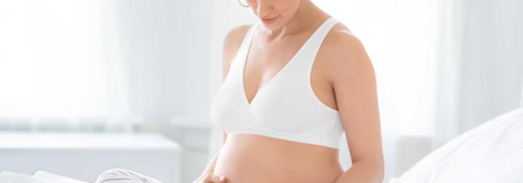 Eine schwangere Frau sitzt in der Mitte ihres Bettes und schaut auf ihren schwangeren Bauch hinunter.