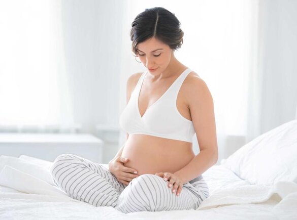 Eine schwangere Frau sitzt in der Mitte ihres Bettes und schaut auf ihren schwangeren Bauch hinunter.