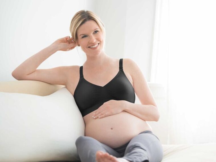Eine schwangere Frau sitzt mit hochgelegten Beinen und der Hand auf dem Bauch auf der Couch und trägt einen Medela-Umstands-BH.