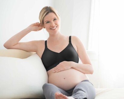 Eine schwangere Frau sitzt mit hochgelegten Beinen und der Hand auf dem Bauch auf der Couch und trägt einen Medela-Umstands-BH.