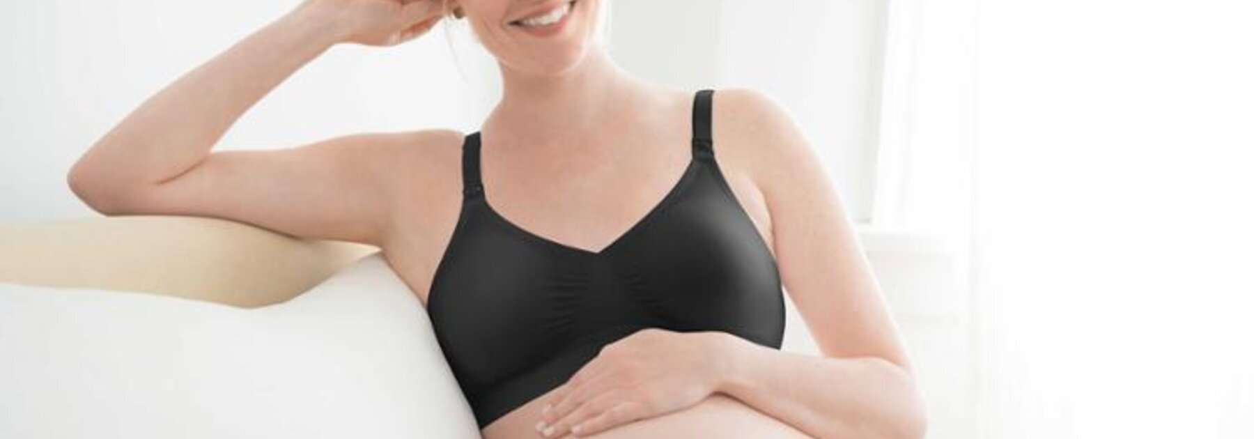 Eine schwangere Frau sitzt mit hochgelegten Beinen und der Hand auf dem Bauch auf der Couch und trägt einen Medela-Umstands-BH.