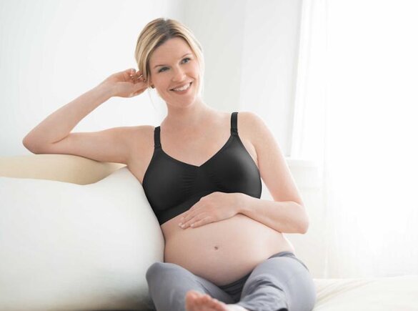 Eine schwangere Frau sitzt mit hochgelegten Beinen und der Hand auf dem Bauch auf der Couch und trägt einen Medela-Umstands-BH.