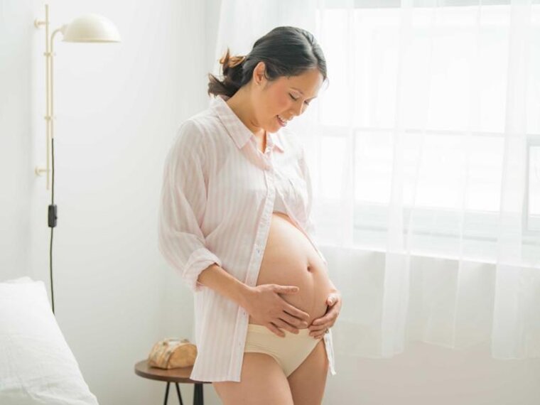 Eine schwangere Frau steht in ihrem Schlafzimmer am Fenster und hält sich ihren unbedeckten schwangeren Bauch.