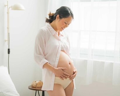 Eine schwangere Frau steht in ihrem Schlafzimmer am Fenster und hält sich ihren unbedeckten schwangeren Bauch.