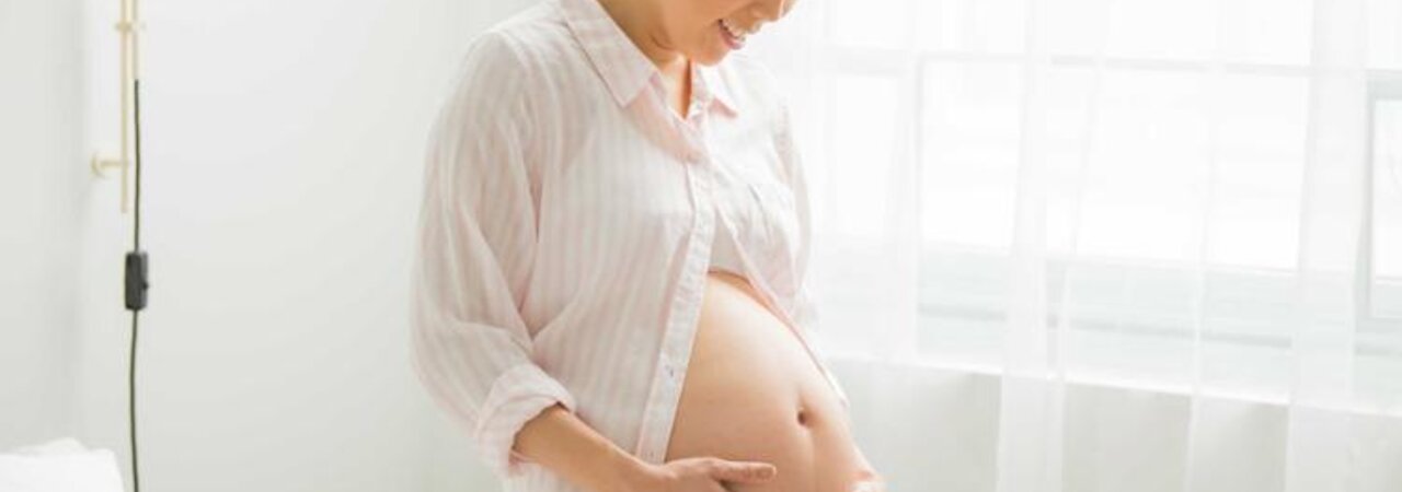 Eine schwangere Frau steht in ihrem Schlafzimmer am Fenster und hält sich ihren unbedeckten schwangeren Bauch.