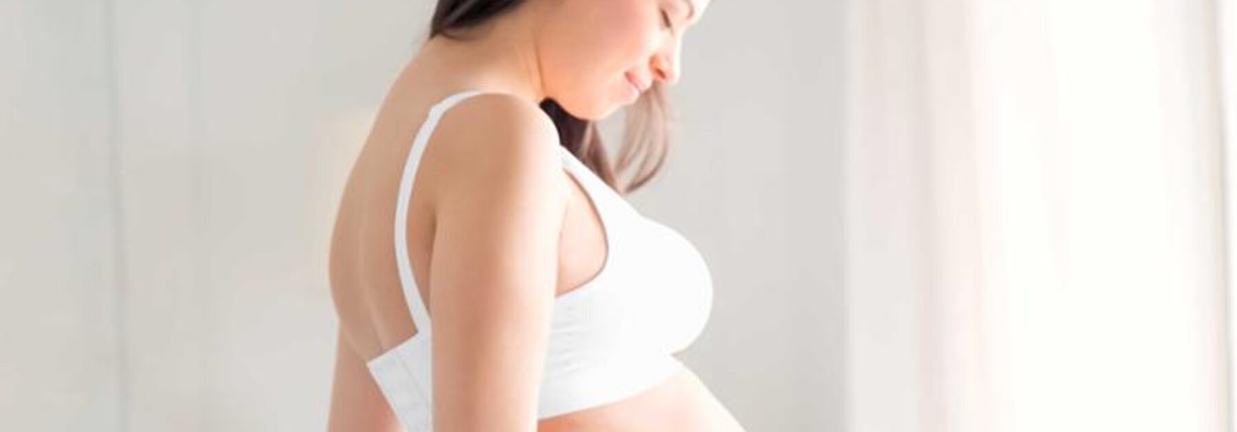 Seitenansicht einer schwangeren Frau, die mit den Händen auf ihrem schwangeren Bauch steht und nach unten schaut.