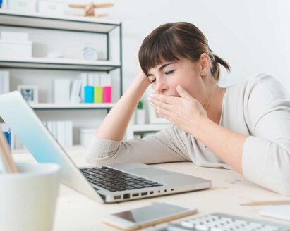 Eine Frau gähnt, während sie an einem Schreibtisch sitzt und an ihrem Laptop arbeitet.