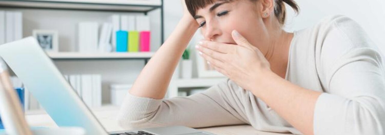Eine Frau gähnt, während sie an einem Schreibtisch sitzt und an ihrem Laptop arbeitet.