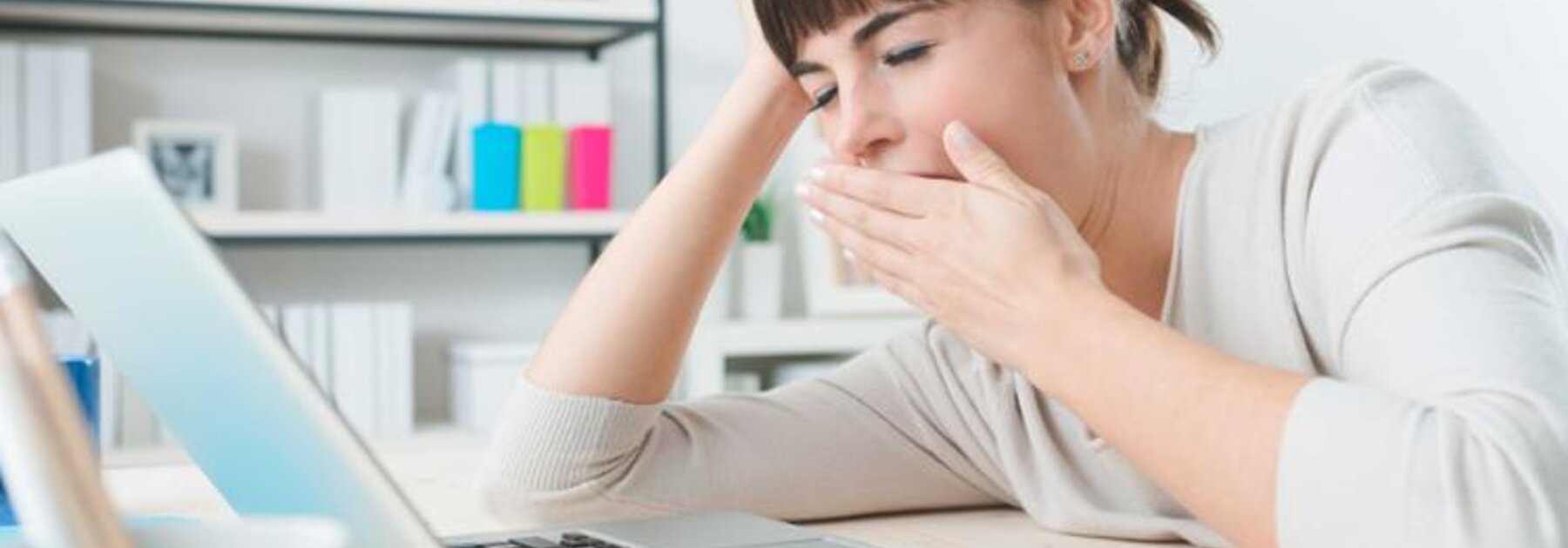 A woman yawns as she sits at a desk on her laptop.