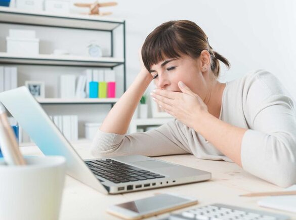 Eine Frau gähnt, während sie an einem Schreibtisch sitzt und an ihrem Laptop arbeitet.