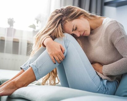 Frau auf einer Couch sitzend, den Kopf auf die Knie gestützt und mit der Hand ihren Bauch haltend.