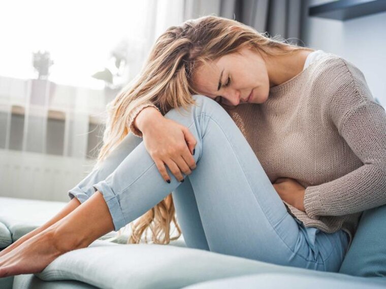 Frau auf einer Couch sitzend, den Kopf auf die Knie gestützt und mit der Hand ihren Bauch haltend.