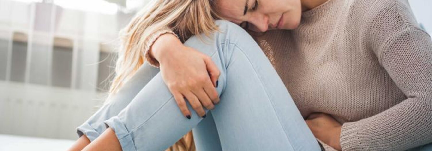 Frau auf einer Couch sitzend, den Kopf auf die Knie gestützt und mit der Hand ihren Bauch haltend.