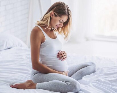 Eine schwangere Frau sitzt in der Mitte ihres Bettes, hat die Beine unter sich angewinkelt und blickt auf ihren schwangeren Bauch.
