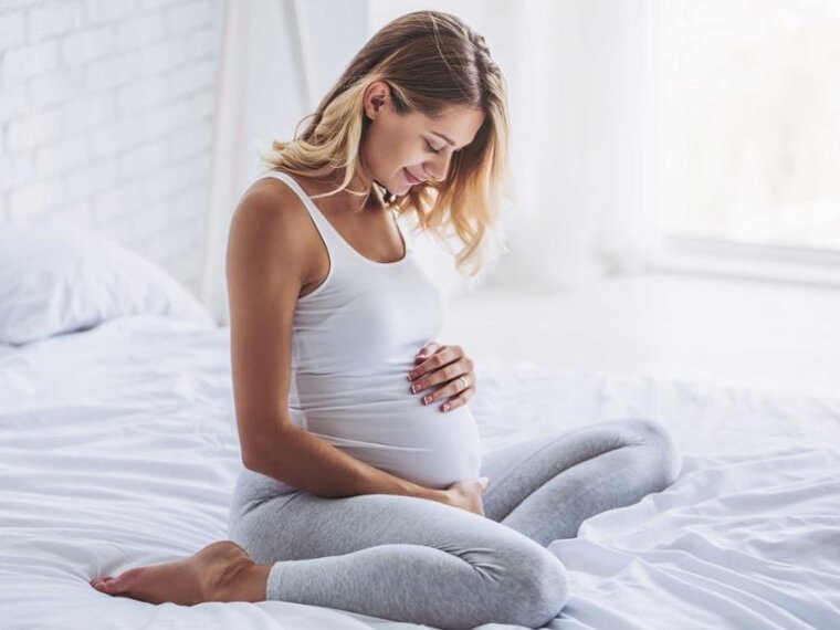 Eine schwangere Frau sitzt in der Mitte ihres Bettes, hat die Beine unter sich angewinkelt und blickt auf ihren schwangeren Bauch.