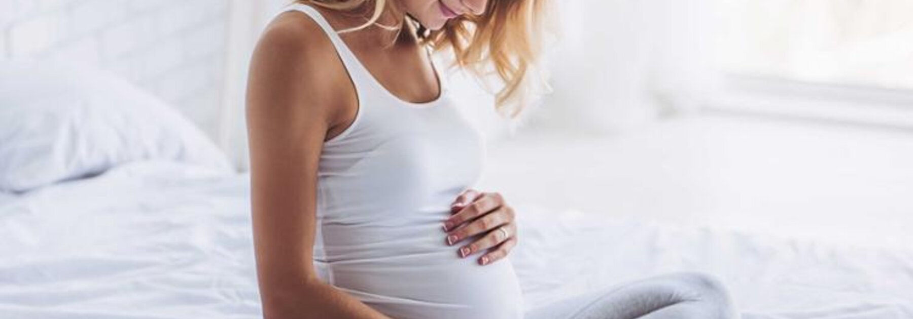 Eine schwangere Frau sitzt in der Mitte ihres Bettes, hat die Beine unter sich angewinkelt und blickt auf ihren schwangeren Bauch.