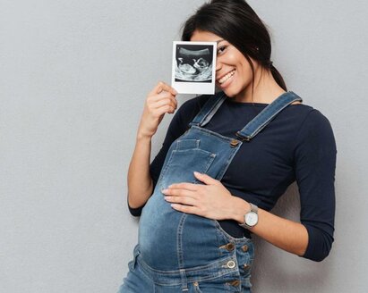 Eine schwangere Frau hält ein Ultraschallbild hoch, während sie ihre Hand auf ihren schwangeren Bauch legt.
