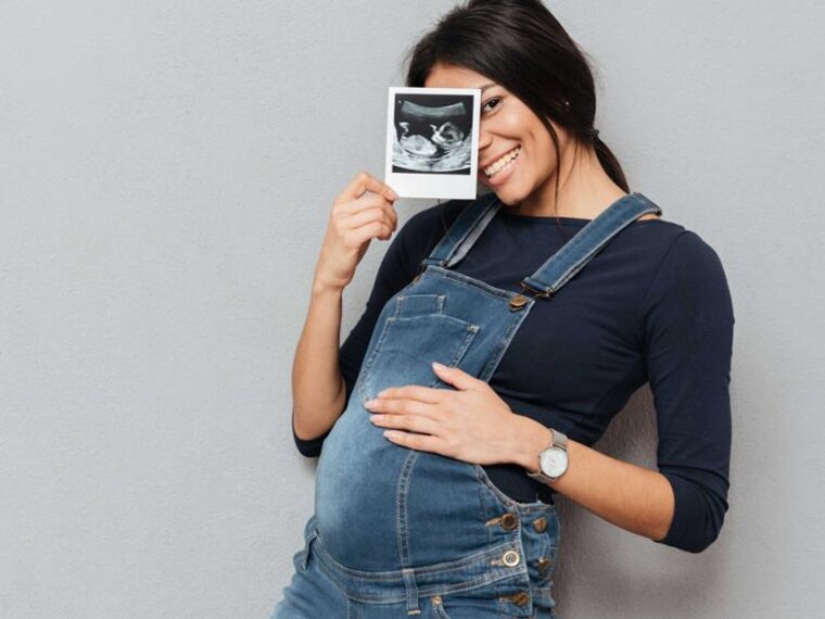 Eine schwangere Frau hält ein Ultraschallbild hoch, während sie ihre Hand auf ihren schwangeren Bauch legt.
