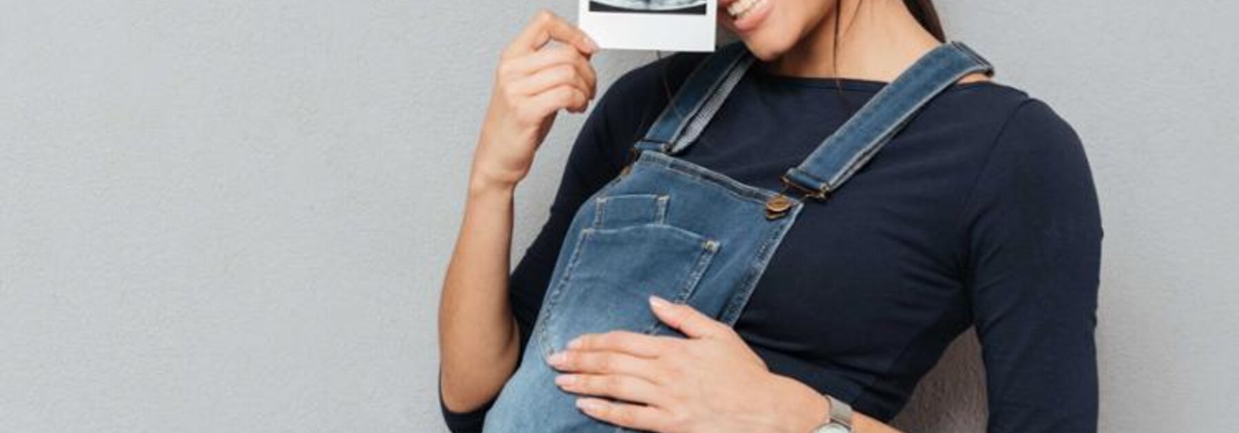 Eine schwangere Frau hält ein Ultraschallbild hoch, während sie ihre Hand auf ihren schwangeren Bauch legt.