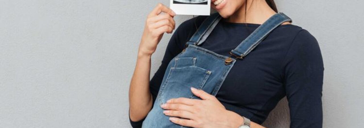Eine schwangere Frau hält ein Ultraschallbild hoch, während sie ihre Hand auf ihren schwangeren Bauch legt.
