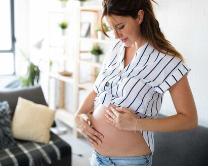 Eine schwangere Frau steht in ihrem Wohnzimmer, legt ihre Hand auf ihren Bauch und spürt, wie sich ihr Baby bewegt.