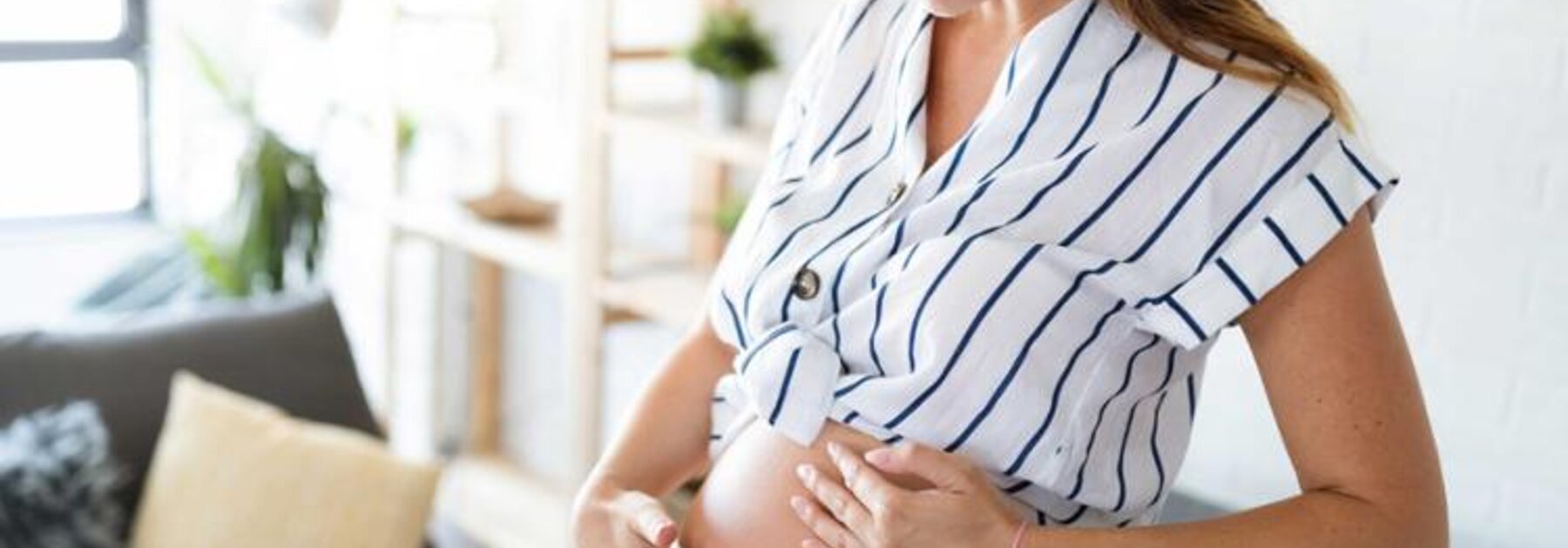 Eine schwangere Frau steht in ihrem Wohnzimmer, legt ihre Hand auf ihren Bauch und spürt, wie sich ihr Baby bewegt.