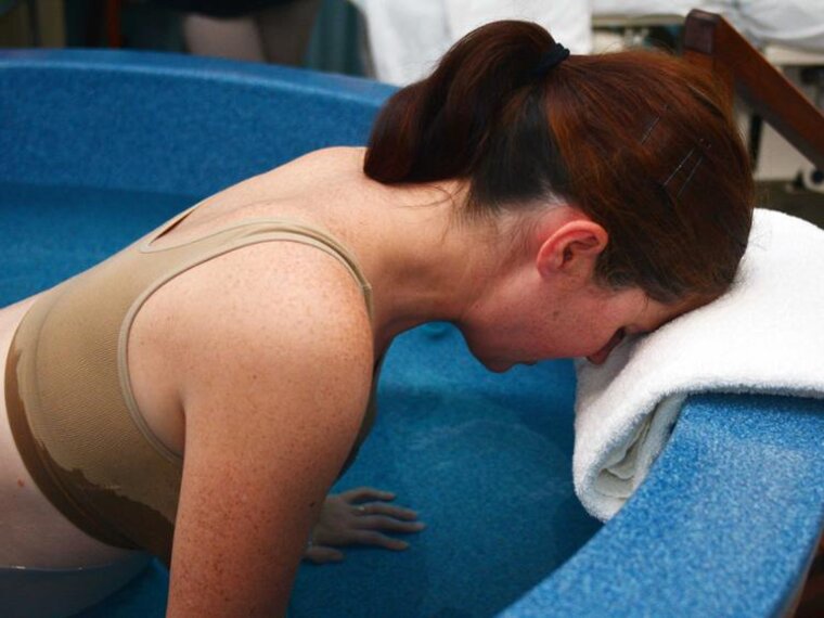 Eine schwangere Frau befindet sich in einem Gebärbecken und kniet mit dem Kopf auf einem Handtuch am Rande.