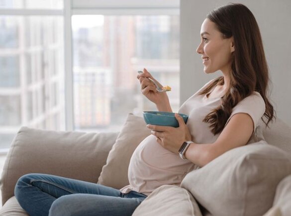 Zwangere moeder eet met haar benen gekruist en kijkt, terwijl ze op de bank zit.