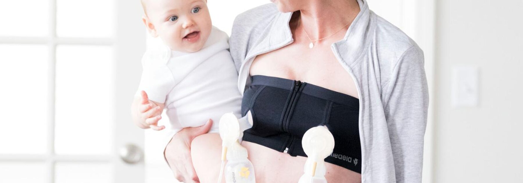 Frau, die ein Medela Easy Expression Bustier trägt, während sie ihr Baby beim doppelten Abpumpen mit einer Medela Milchpumpe hält.