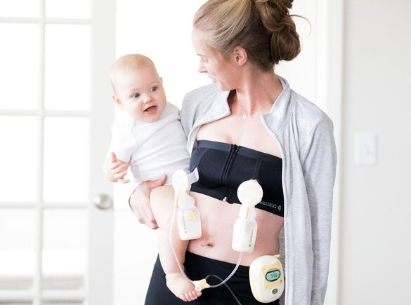 Frau, die ein Medela Easy Expression Bustier trägt, während sie ihr Baby beim doppelten Abpumpen mit einer Medela Milchpumpe hält.