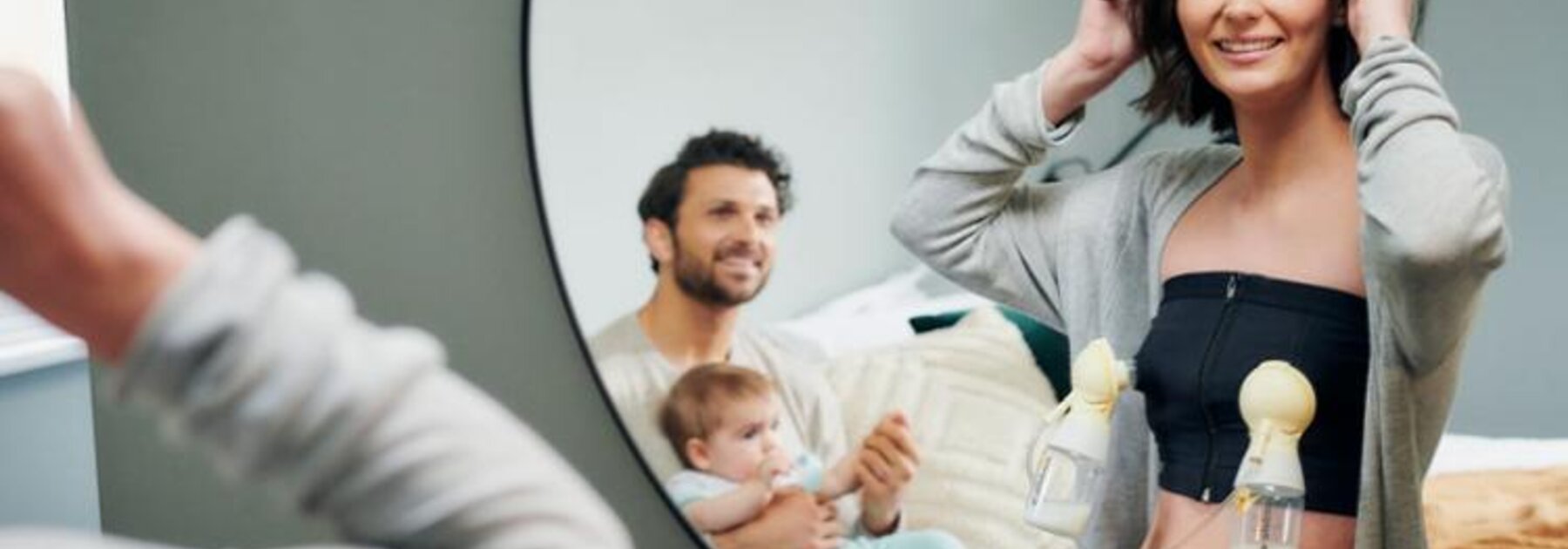 Una madre se extrae leche con un sacaleches manos libres Medela mientras se prepara para ir a trabajar, mientras el padre y el bebé observan desde la otra habitación.