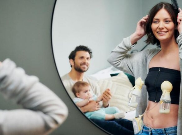 Una madre se extrae leche con un sacaleches manos libres Medela mientras se prepara para ir a trabajar, mientras el padre y el bebé observan desde la otra habitación.